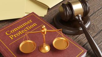 Golden scales of justice on a brown leather bound book engraved with the title Consumer Protection, together with a gavel and yellow office folders on a wooden table